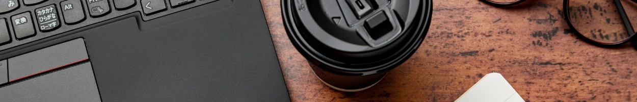 木目調のデスクの上に置かれたノートパソコン、テイクアウト用のコーヒーカップ、眼鏡、そしてノートの角が見える俯瞰写真。