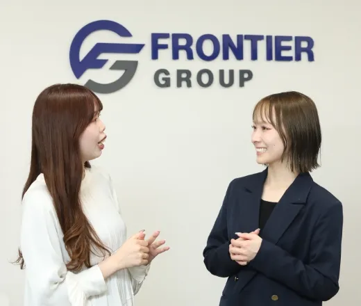 FRONTIER GROUPのロゴの前で楽しそうに会話をする2人の女性社員。