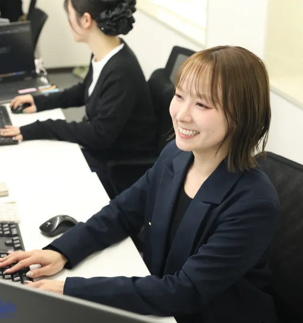 オフィスでパソコンに向かって笑顔で作業をする、紺色のジャケットを着た女性社員。