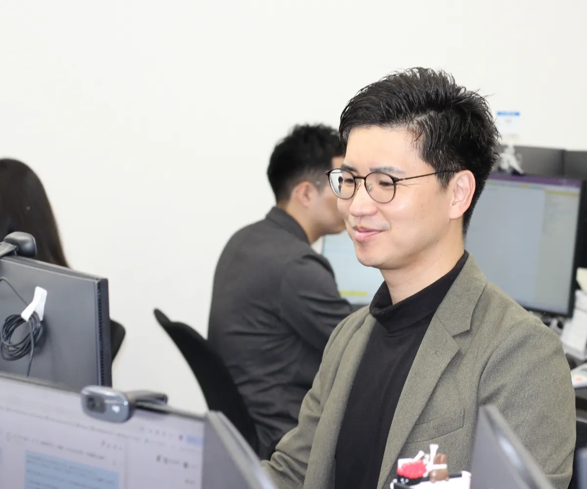 眼鏡をかけ、オフィスでパソコンを操作しながら同僚と談笑する男性社員。